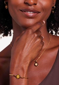 Gouden armband met een ronde gele steen, gecombineerd met een delicate gouden ketting met een harthanger. Textuurdetails van de huid zichtbaar.