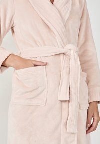 Mjukt rosa fleecebadrock med sjalkrage, knytbälte och fickor med lappar. Har en plyschig textur och fullängdsärmar.
