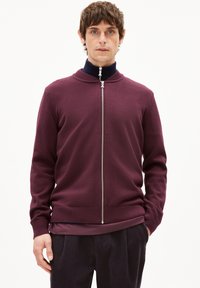 Pull-over bordeaux à fermeture éclair avec un col haut, fabriqué dans un matériau en maille lisse. Il présente une fermeture éclair argentée et un ourlet côtelé, superposé sur un haut sombre.