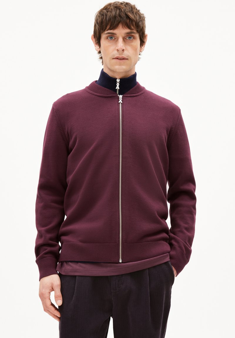 Pull-over bordeaux à fermeture éclair avec un col haut, fabriqué dans un matériau en maille lisse. Il présente une fermeture éclair argentée et un ourlet côtelé, superposé sur un haut sombre.