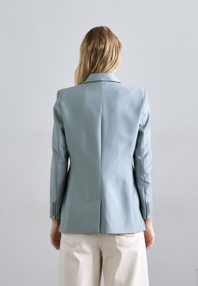 Blondyn stojący twarzą do ściany, w jasnoniebieskim dopasowanym żakiecie i kremowych spodniach z dekoracyjnym przeszyciem na tylnych kieszeniach.