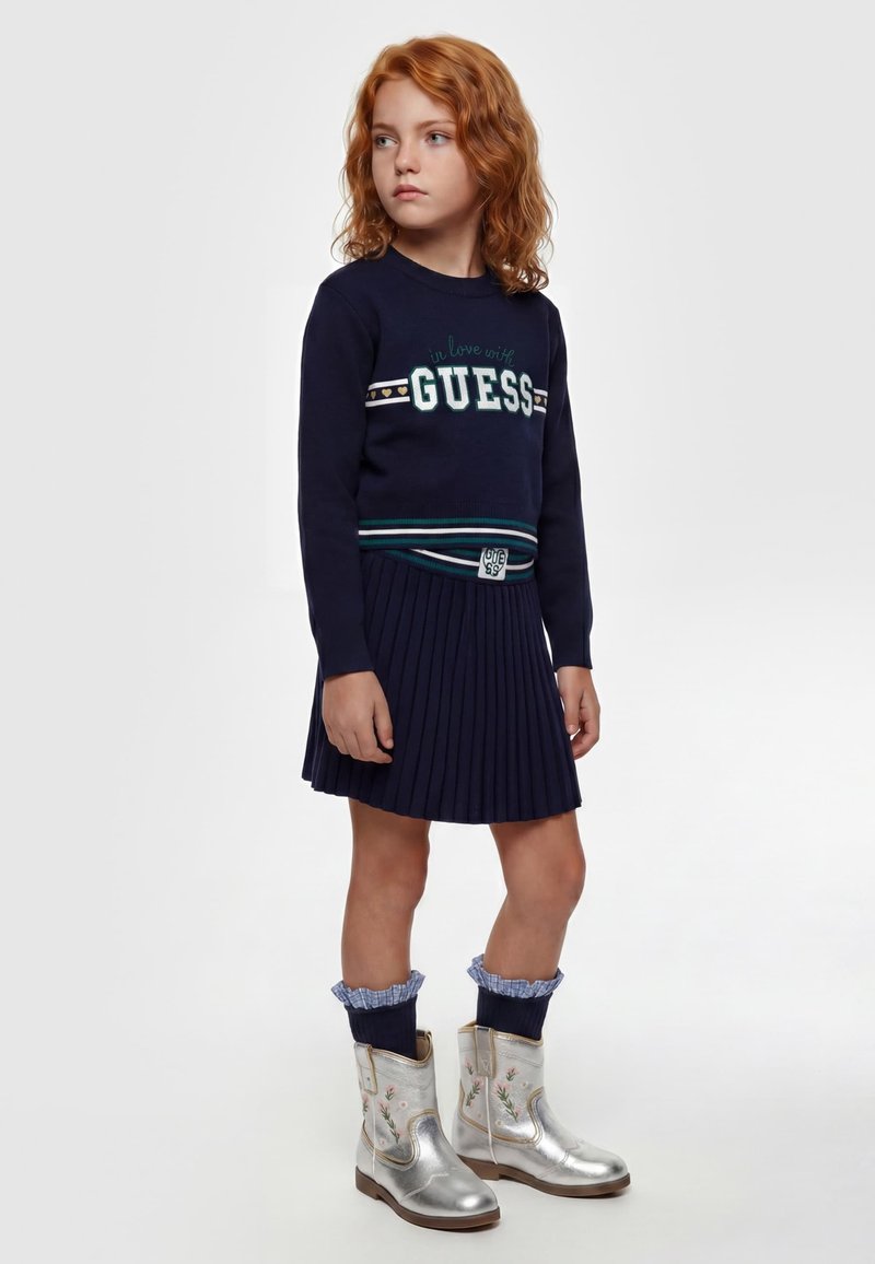 Jeune fille aux cheveux roux portant une robe-pull bleu marine et des bottes de cowboy argentées à motif floral, debout et regardant sur le côté sur un fond uni.