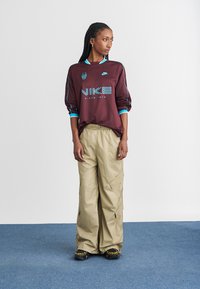 Camisa de manga longa da Nike em bordeaux com detalhes e logotipo em turquesa; combinada com calças cargo folgadas em bege com múlticos bolsos. Calçado preto.