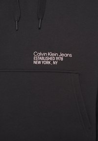 Felpa con cappuccio nera, con cordini e tasca frontale. Presenta un testo bianco: "Calvin Klein Jeans FONDATO NEL 1978 NEW YORK, NY."