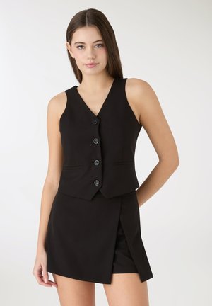 Gilet nero su misura con sei bottoni, abbinato a una gonna nera con spacco frontale. Tessuto liscio e design aderente.