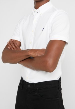 Persona que lleva una camisa blanca de manga corta con un pequeño logo y pantalones negros, con los brazos cruzados sobre el pecho.