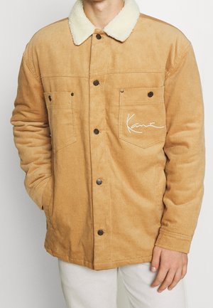 Hombre con chaqueta de pana color beige con cuello de sherpa blanco, cierre de botones, dos bolsillos en el pecho y firma bordada en blanco en el bolsillo derecho.