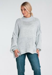 Jasnoszary, oversize'owy sweter dziany z wysokim kołnierzem, szerokimi rękawami i teksturowanym wzorem. Zestawiony z niebieskimi dżinsami.