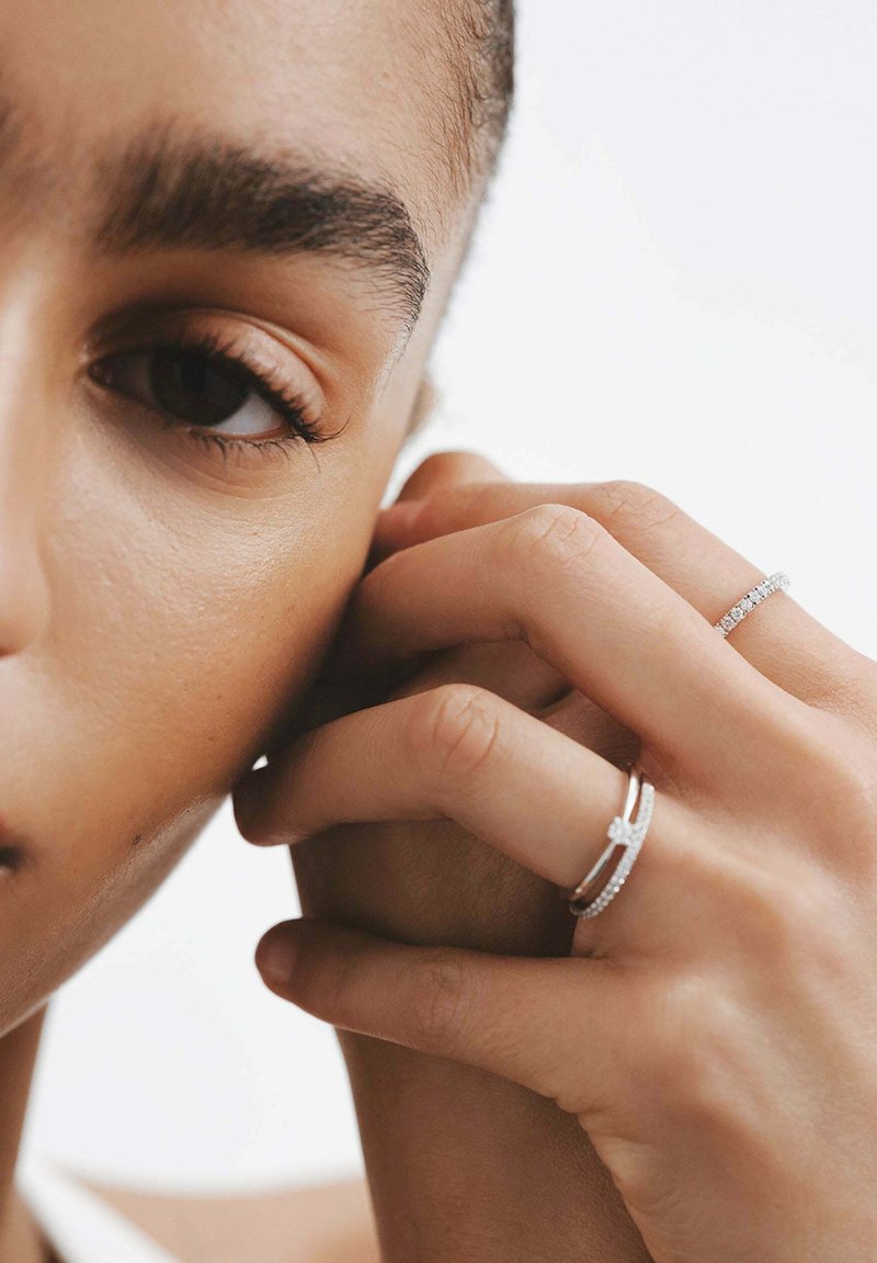 Se llevan anillos de plata en los dedos, con un diseño delgado y uno con pequeñas piedras incrustadas. Primer plano de una oreja y una mano. Textura de piel suave.