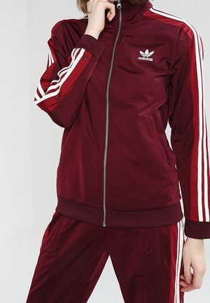 Chaqueta deportiva burdeos con rayas blancas en las mangas, cremalleras en el frente y un pequeño logotipo blanco. Textura suave con un diseño ajustado.