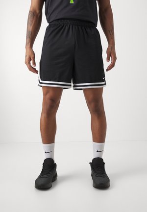 Mann trägt schwarze Basketballshorts mit weißen Streifen, schwarze Sneakers und weiße Nike-Socken, steht auf einem weißen Boden.