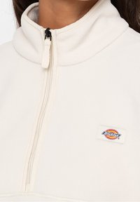 Maglione in pile crema con colletto alto e zip a un quarto. Presenta una zip nera e un colorato patch del logo Dickies sul petto.