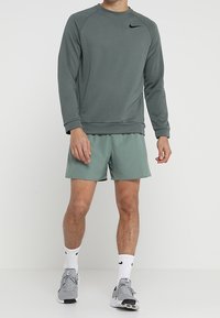 Bluza Nike w stonowanej zieleni, z okrągłym dekoltem i prążkowanymi mankietami. Połączona z jaszielonymi szortami i szarymi butami sportowymi.