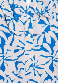 Tessuto bianco con motivo floreale blu, collo a camicia e patta con bottoni. I dettagli includono accenti arricciati al collo e una texture liscia.
