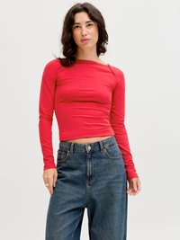 Kvinne med mørkt bølget hår iført en tettsittende rød crop-top med lange ermer og løstsittende blå jeans med høy midje, stående foran en hvit bakgrunn.