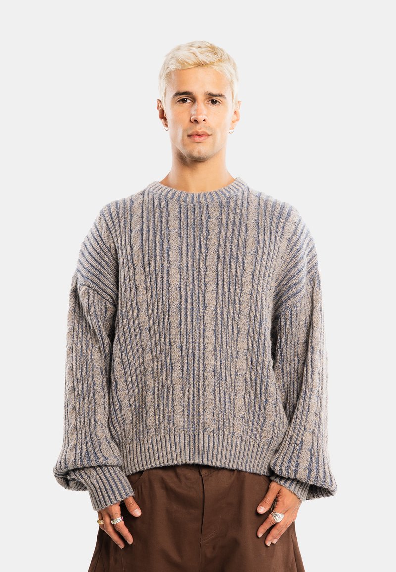 Jeune homme aux cheveux blonds courts portant un pull en maille torsadée ample gris et bleu et un pantalon marron, debout devant un fond uni.