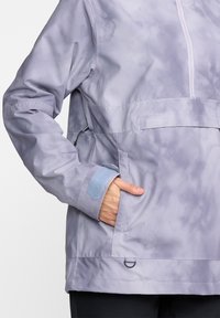 Chaqueta impermeable de color púrpura claro con acabado brillante, cremallera frontal, bolsillos laterales y puños de velcro azules en contraste. Cuenta con un ajuste en el dobladillo.