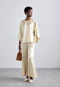 Light beige satin top with a keyhole neckline and oversized sleeves paired with matching flared trousers. Brown perforated handbag and pointed heels.