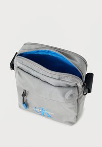 Γκρι υφασμάτινη τσάντα χιαστί με μπλε εσωτερικό, κλείσιμο με φερμουάρ, μπροστινή τσέπη και κεντημένο λογότυπο "CK" σε μπλε χρώμα στο μπροστινό μέρος.