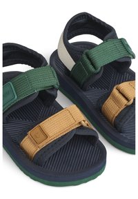 Δύο σανδάλια με ναυτικό μπλε βάση· πράσινα και καφέ λουριά από υφασμένο υλικό με υφή. Διαθέτουν εύκαμπτη σόλα με ραβδώσεις για καλύτερο κράτημα.