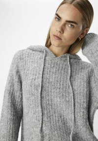 Grå stickad huva med ett strukturellt mönster, snöre och lös passform, tillverkad av mjukt, fluffigt material. Betonar en avslappnad design.