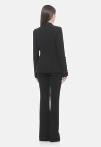 Blazer nero su misura con vita aderente, maniche lunghe con dettagli di bottoni, abbinato a pantaloni neri svasati. Tessuto dalla texture liscia.