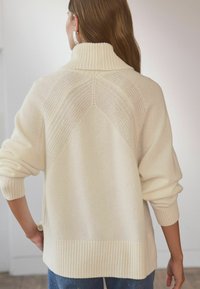 Cremefarvet strikket sweater med høj krave, der har ribbede detaljer og et tekstureret rygdesign. Løs pasform med dropped skuldre.