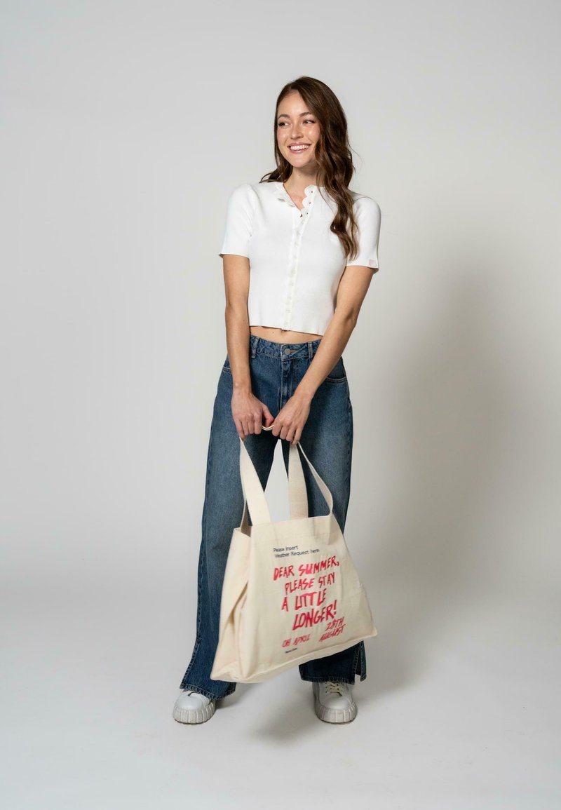 Sac en toile beige clair avec du texte rouge. Modèle portant un top court blanc et un jean large bleu, se tenant avec le sac.