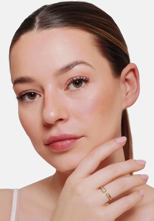 Kobieta z gładką skórą i brązowymi oczami delikatnie dotykająca swojej twarzy, nosząca złoty pierścionek z dwoma kamieniami na palcu.