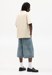 Beige oversized polo skjorte, lys blå denim culotter med lodrette striber, sorte snørebåndssko og hvide ankelsokker, set fra ryggen.