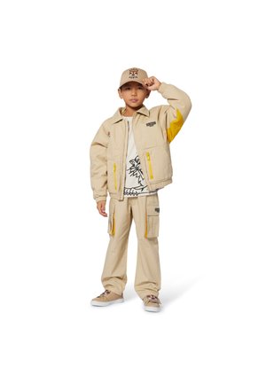 Beige cargojack met gele accenten, bijpassende cargobroek en een graphic t-shirt. Een beige pet met logo. Casual schoeisel maakt de look af.