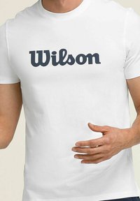 Camisa blanca de manga corta con el logo "Wilson" en azul en el pecho, llevada por una persona con una mano ligeramente levantada y relajada.