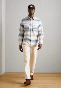 Multi-farbiges kariertes Flanellhemd mit zwei Fronttaschen, cremefarbene Hose und brauner Mütze, kombiniert mit braunen Schuhen auf einem Holzfußboden.