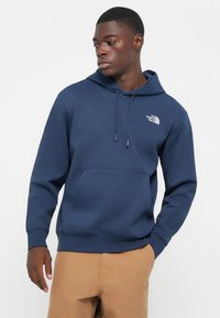 Marineblauwe hoodie gemaakt van fleece materiaal. Voorzien van een voorzak en een wit logo op de linkerkant van de borst. Ribgebreide manchetten en zoom.