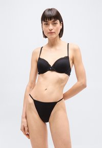 Γυναίκα με κοντά σκούρα μαλλιά που φοράει μαύρο σουτιέν με δαντέλα και ταιριαστά σλιπ με ψηλή μέση, στέκεται μπροστά σε απλό φωτεινό φόντο.