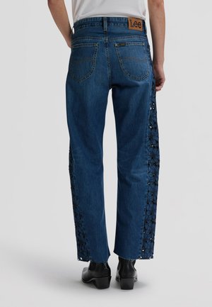 Άτομο που φορά μπλε τζιν με floral δαντελένιες επενδύσεις στα πλάγια, μαύρα μποτάκια αστραγάλου και λευκό μπλουζάκι, εμφανιζόμενο από πίσω.