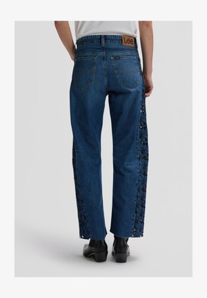 Oseba, ki nosi modre kavbojke iz jeansa z cvetličnim čipkastim vložkom na straneh, črne gležnjarje in belo majico, prikazana od zadaj.