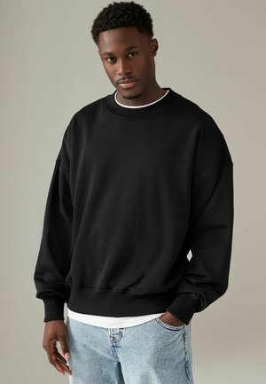Svart oversized sweatshirt med ribbad halsringning och muddar, i kombination med en vit T-shirt under och ljusblå jeans. Slät textur.