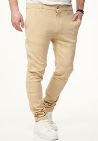Beige enganierte Hose aus einem glatten Baumwoll-Mix-Gewebe. Mit einem vorderen Knopfverschluss, Seitentaschen und minimaler Nahtdetailierung.