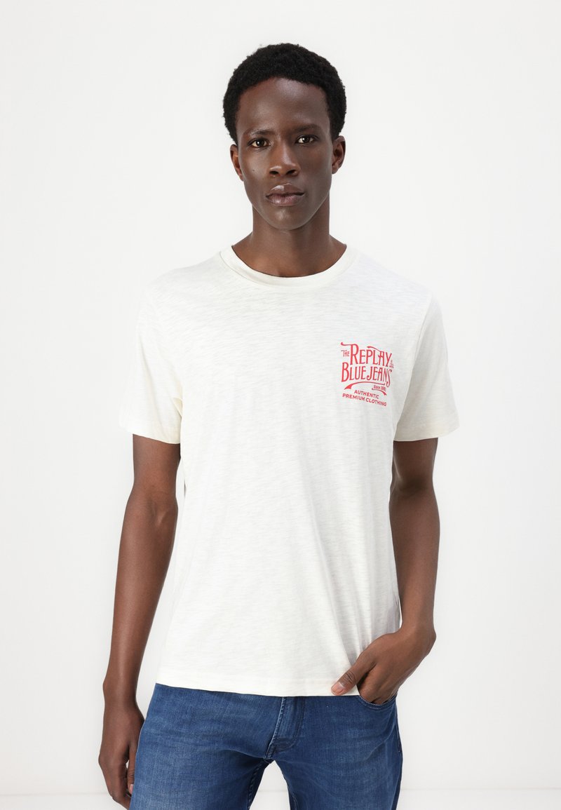 Helles Creme-Kurzarm-T-Shirt mit Rundhalsausschnitt. Mit roten Textgrafiken auf der linken Brust. Hergestellt aus einem weichen, strukturierten Stoff.