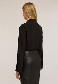 Blusa nera con maniche plissettate, indossata con una gonna a tubino in pelle nera. Il tessuto liscio e il design sartoriale creano una silhouette elegante.