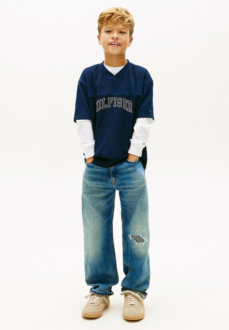 Młody chłopiec stoi z rękami w kieszeniach, ubrany w warstwowe granatowo-białe koszulki z długim rękawem, podarte niebieskie dżinsy i beżowe trampki na białym tle.