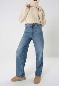 Pull à col roulé beige en maille avec une texture côtelée. Jean taille haute bleu clair à jambes larges. Bottines cheville en daim beige.