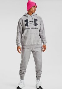 Felpa grigia con grande logo nero, tasca frontale e cordini. Pantaloni della tuta grigi con caviglie elastiche. Berretto fucsia e sneakers bianche.