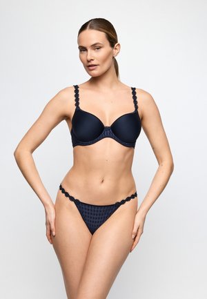 Kobieta stojąca w granatowym komplecie bielizny z kwiatowymi ramiączkami i teksturowanymi majtkami bikini na białym tle.
