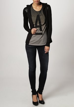 Sort lynlåsjakke med læreærmer, grå T-shirt med stort bogstav A, mørke skinny jeans og sorte højhælede sko.