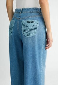 Jeans a vita alta in denim con tasche posteriori in crochet blu chiaro con un motivo geometrico e lettering ricamato. Vestibilità rilassata con gambe dritte.