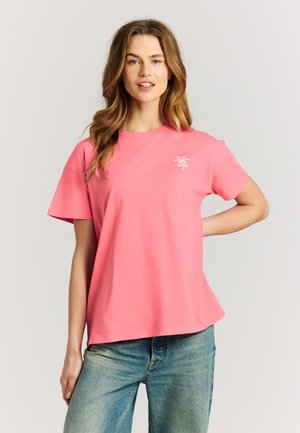 Jonge vrouw met lang haar die een roze T-shirt draagt met een kleine witte palmboom en tekst op de borst, gecombineerd met een blauwe spijkerbroek, poserend met hand op heup.