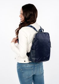Mujer con cabello largo y oscuro, que lleva una chaqueta blanca y jeans azules, con una mochila azul marino en la espalda, sonriendo y mirando hacia la izquierda.