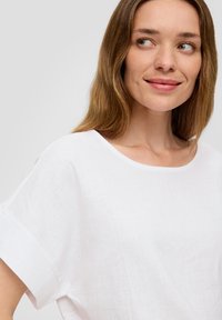 Blouse en lin blanc avec un large décolleté et des manches courtes retournées, présentant une texture douce et une coupe ample pour le confort.
